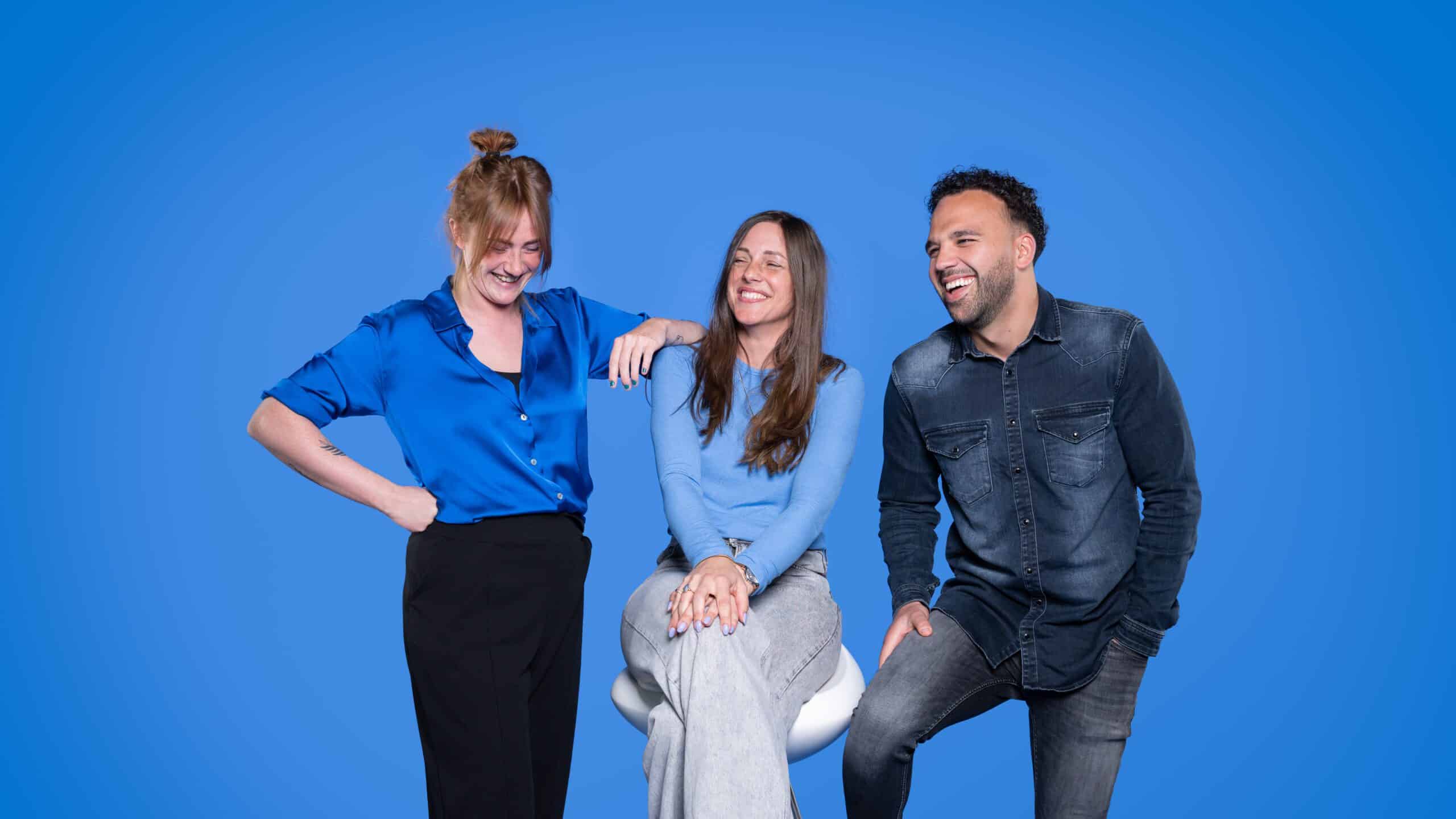Teamfoto van drie collega’s lachend voor een helderblauwe achtergrond